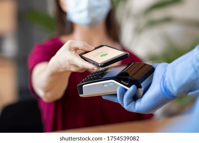 Cashier hand holding credit card reader machine and wearing disposable gloves while client holding phone for NFC payment. Woman wearing face mask while paying with smartphone during Covid-19 pandemic.