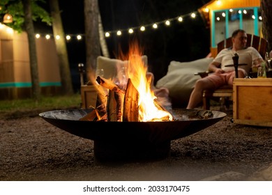 Cast iron fire pit campfire place at forest beach camping with brgiht burning flame at evening time against light bulb garland and trees. Friends sit near camp bonfire stove at warm summer night