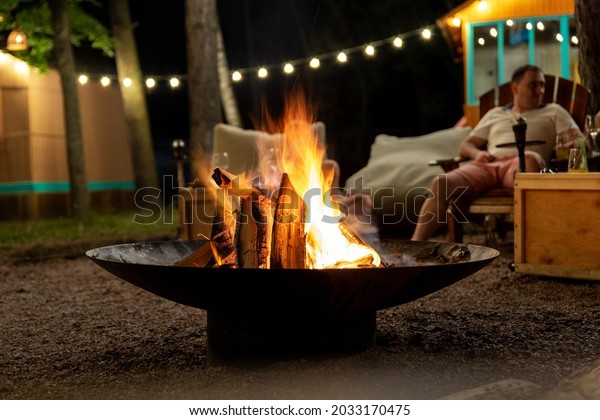 Cast iron fire pit campfire place at forest beach\
camping with brgiht burning flame at evening time against light\
bulb garland and trees. Friends sit near camp bonfire stove at warm\
summer night
