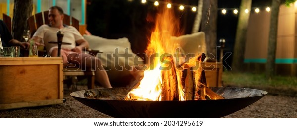 Cast iron fire pit campfire place at forest beach\
camping with brgiht burning flame at evening time against light\
bulb garland and trees. Friends sit near camp bonfire stove at warm\
summer night