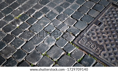 Cast iron manhole cover with SPQR engraving on a stone floor with a geometric design in the morning sun