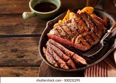Cast iron plate with sliced grilled rib-eye steak on table with yellow potato wedges and oil in saucer over table with copy space