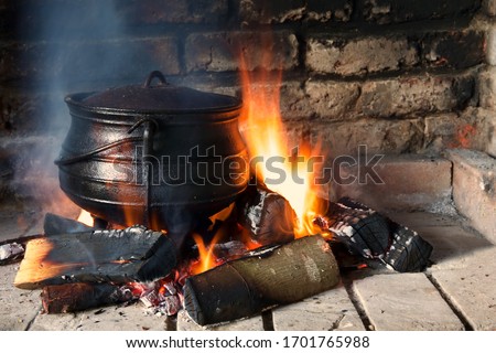 Cast iron three legged pot on a wooden fire in a brick fire place.  Smoke and flame surround the pot.
