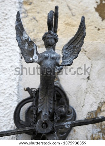 Cast iron winged angel figure atop a bell set against blurred cracked old wall background in southern France