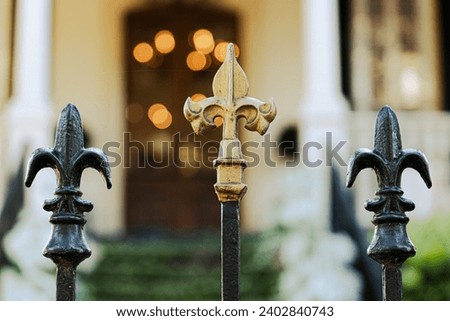 A cast wrought iron fence lined with black and gold fleur de lis post toppers with a New Orleans southern style home in the background.