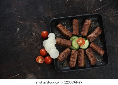 Cast-iron serving pan with grilled cevapi or cevapcici sausages, flatlay with copyspace on a dark brown stone background