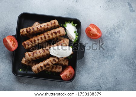 Cast-iron serving tray with grilled cevapi or cevapcici sausages and kajmak cheese, above view on a light-blue stone background, copyspace