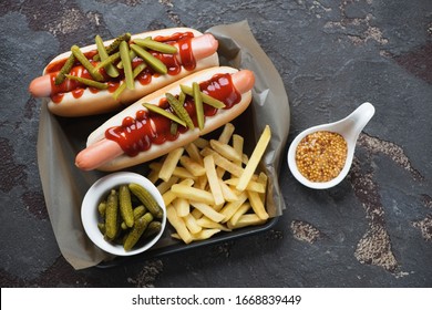 Cast-iron serving tray with two hot-dogs, french fries and pickles, high angle view over brown stone background, studio shot