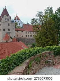 Castle structures with red roofs stand tall among green trees. A stone path winds along the wall, leading to a view of the buildings. Clouds hang overhead, adding to the atmosphere.