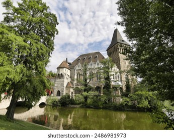 Castle-like building with turrets and a bridge over a tranquil pond, surrounded by lush trees and greenery, under a partly cloudy sky. - Powered by Shutterstock