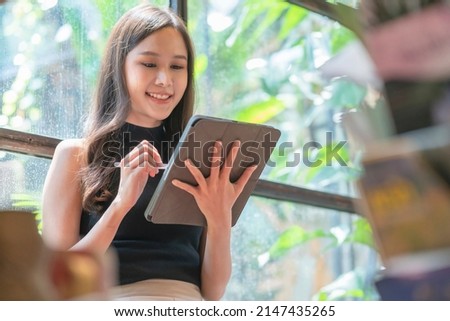 casual asia female woman relax hand using tablet internet surf writing diary cheerful smiling while sittting in warm interior of cafe near big window,asian woman sit on stool casual work in coffeeshop