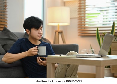 Casual Asian Male Drinking Coffee And Reading Online News On His Laptop While Sitting In Cozy Living Room