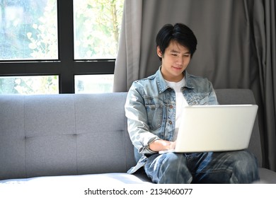 Casual Asian Man Resting On Couch At Home And Browsing Internet With Computer Laptop.