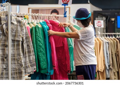 Casual Asian woman wears beanie with protective mask choosing jacket clothes on clothing rack in fashion store at shopping mall - Powered by Shutterstock