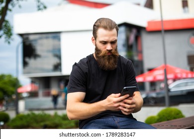 Casual Attractive Man With A Beard Relaxing On A Park Bench Reading A Tex Message On His Mobile Phone