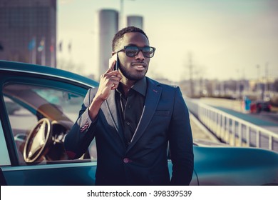 Casual black man in a suit talking by smartphone near a car.