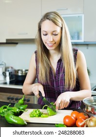 Casual blonde young woman slicing green pepper at table at domestic kitchen