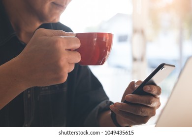 Casual Business Man, Freelancer Using Mobile Smart Phone, Drinking Coffee, Working On Laptop Computer Near The Window At Home Office With Morning Sunlight, Close Up