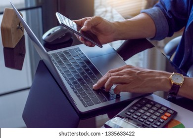 Casual business man hand using mobile smart phone and typing on laptop computer keyboard with calculator and data graph paper on the mirror table. Digital marketing, ecommerce concept.