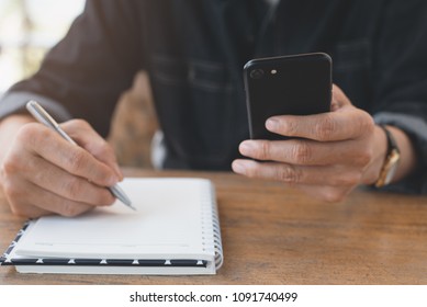 Casual Business Man Using Mobile Smart Phone Working, Taking Note, Schedule On Paper Notebook, Reading E Book Application, Searching On Internet For Research In Coffee Shop. E Learning Concept