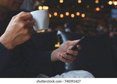 Casual Business Man Using Mobile Smart Phone And Drinking Coffee In Coffee Shop Or Cafe At Night With Dark Background And Colorful Bokeh, Internet Network Communication, Modern Lifestyle Concept.