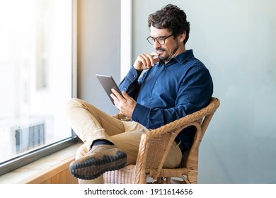 Casual Business Man Wearing Glasses Sitting Looking At His Tablet. Successful Male Portrait With Thinking Face Expression Working From Home By The Window