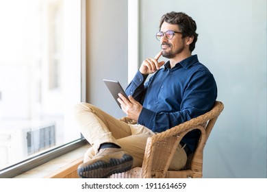Casual business man wearing glasses sitting holding his tablet. Successful male portrait with thinking face expression working from home by the window