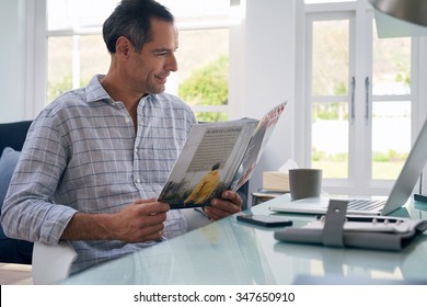 Casual Businessman Seated At Home Office Space Reading Business Magazine Smiling