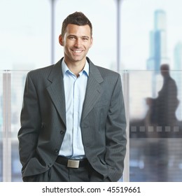 Casual Businessman Standing With Hands In Pocket In Corporate Office Lobby, Smiling.