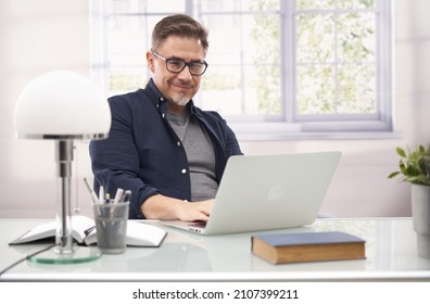 Geschäftsmann, der mit Laptop-Computer zu Hause arbeitet, glücklich, lächelnd. Porträt des mittleren Alters in den 50ern, weißes, kaukasisches, graues Haar.
