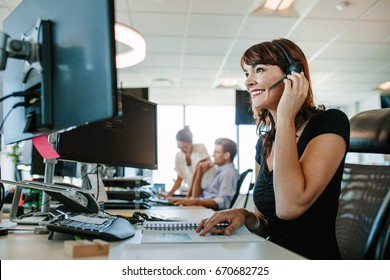 Casual Businesswoman Working At Desk Using Computer And Headset In The Office. Focus On Woman Sitting In Foreground With Colleagues Working In Background.