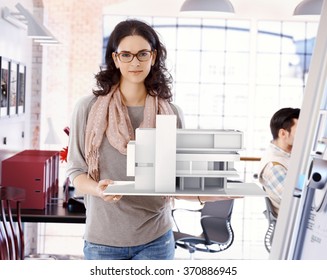 Casual Caucasian Attractive Female Architect With Scale Model Of A Building Plan, Standing, Holding, Wearing Glasses, Looking At Camera, Smiling At Office.