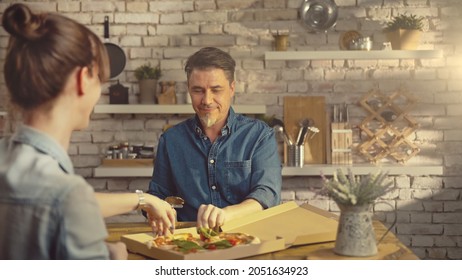 Casual Couple Eating Take Away Pizza From Food Box. Man Sharing Pizza On Kitchen Table At Home.