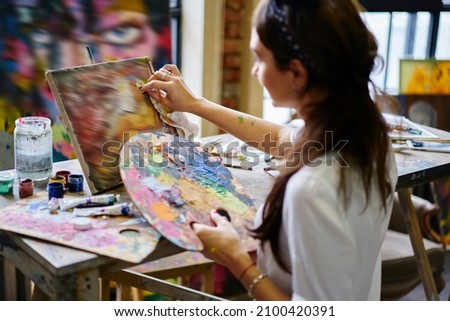 Casual dressed female artist with palette for mixing acrylic colors spending daytime for art workshop in studio, Caucasian woman inspired during masterpiece artwork in gallery drawing picture indoors