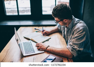 Casual dressed hipster guy writing organisation plan in textbook for education sitting at desktop with netbook,young man writing essay in notebook and preparing for academy test with modern technology