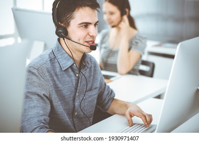 Casual dressed young man using headset and computer while talking with customers online. Group of operators at work. Call center - Shutterstock ID 1930971407
