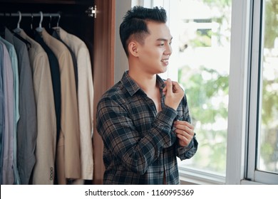 Casual Handsome Asian Man Wearing Checkered Shirt At Home Getting Dressed Near Wardrobe And Looking Away With Smile