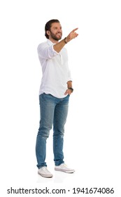 Casual Happy Man In White Shirt, Jeans And Sneakers Is Standing With Hand In Pocket, Looking Away And Pointing. Front Side View. Full Length Studio Shot Isolated On White.