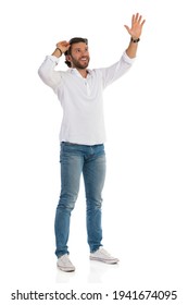 Casual Happy Man In White Shirt, Jeans And Sneakers Is Standing, Looking Up, Holding Hand Raised And Waving. Front Side View. Full Length Studio Shot Isolated On White.