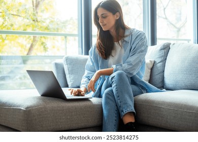 Casual home scene of a young woman in denim, sitting on her sofa with a laptop, multitasking between remote work, job hunting, and online chatting, enjoying the benefits of wireless - Powered by Shutterstock