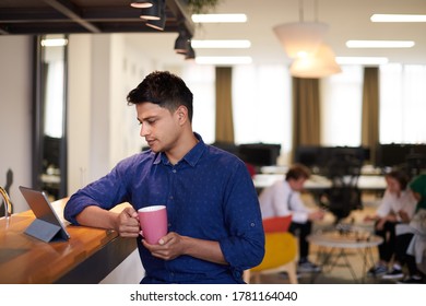 Casual Indian Business Man Taking Break From The Work  Using Tablet Computer While Drinking Tea In Relaxation Area Of Modern Open Plan Startup Office