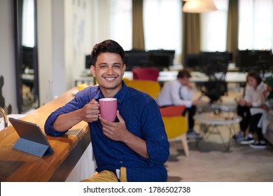 Casual Indian Business Man Taking Break From The Work  Using Tablet Computer While Drinking Tea In Relaxation Area Of Modern Open Plan Startup Office