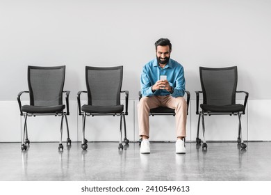 Casual indian businessman seated alone, absorbed in his smartphone, in stark waiting area with rows of empty chairs, suggesting moment of downtime
