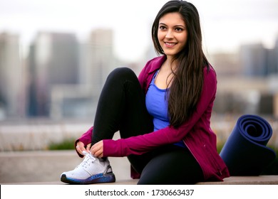 Casual lifestyle portrait of beautiful Indian American woman relaxing after cardio fitness exercise, city skyline buildings in background