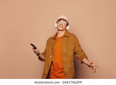 Casual man in a denim jacket stands against a peach studio background. He smiles, holding a mobile phone and wearing glasses, immersed in his music. He dances happily to his favorite song. - Powered by Shutterstock