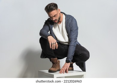 Casual Man With Eyeglasses Looking Down While Holding Elbow On Knee And Crouching In Studio 