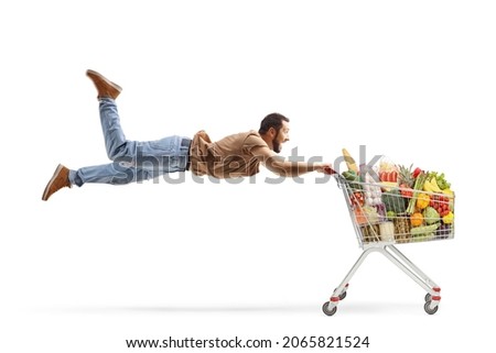 Casual man flying and holding onto a shopping cart filled with food products isolated on white background