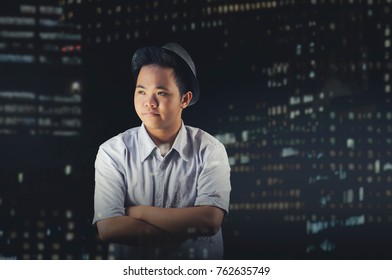 Casual Man Wearing A Vintage Leather Black Hat, Standing And Crossed Arms Behind An Office Window Glass With Skyline Bokeh Background From Building Lights. No Emotions Or Thinking About Something.