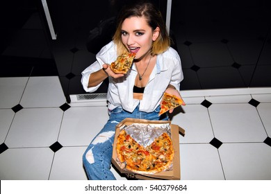 Casual portrait of amazing blonde woman sitting on floor at home, holding two pieces of hot tasty delicious pizza. Pretty girl wearing white stylish shirt and trendy jeans. 