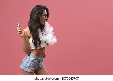 Casual pretty woman standing and vaping on pink background in studio.